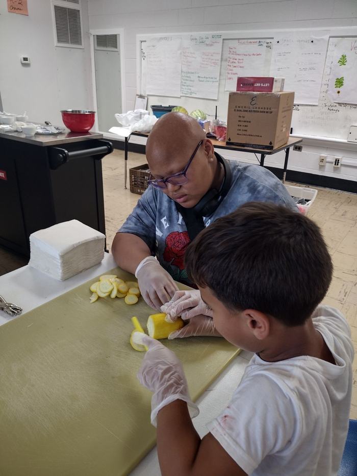 Adult supervising child slicing yellow squash on cutting board, both wearing gloves.