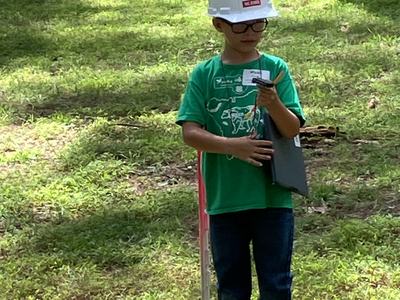 Child wearing white hard hat and glasses holding clipboard and measuring device in grass