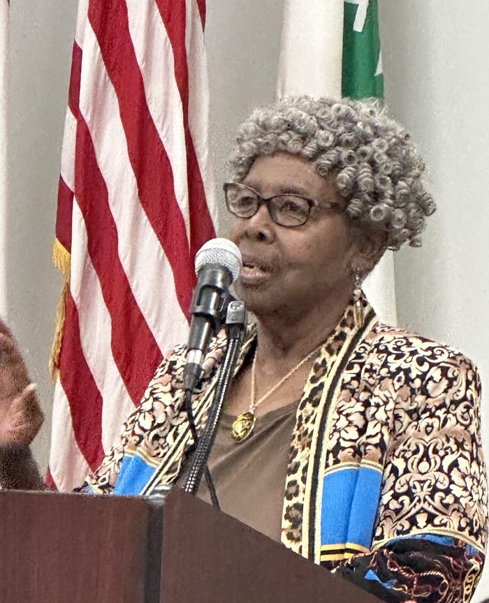 Older woman speaking at a podium into a microphone with a U.S. flag behind
