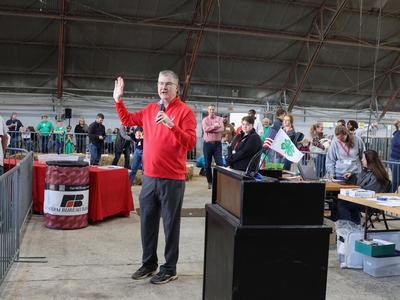 Man in red sweater holding a microphone and raising his hand while speaking at an indoor fair
