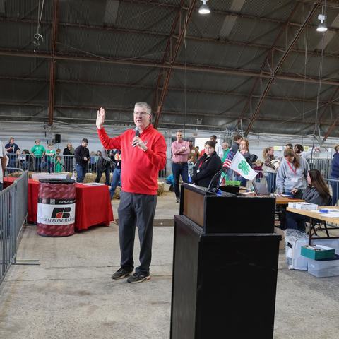 Man in red sweater holding a microphone and raising his hand while speaking at an indoor fair