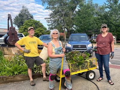 Three gardeners standing by a yellow cart of pulled plants and tools, one shirt reads "Stop Litter"