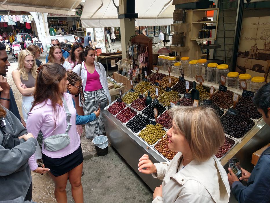 Group of people sampling and buying olives at an outdoor market stall