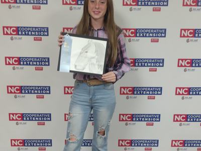 Teen girl holding framed horse drawing with blue ribbon in front of NC Cooperative Extension backdrop