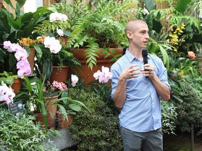 Man holding a microphone speaking in a greenhouse among orchids; sign reads "Please do not touch"