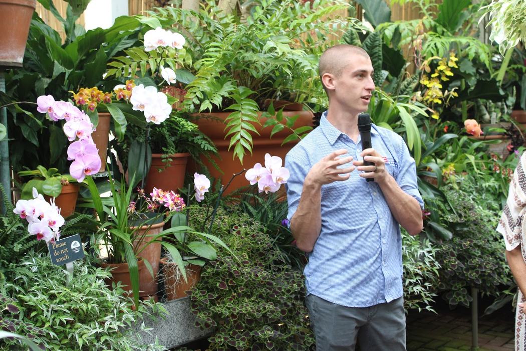 A man speaks in front of a display of potted plants.