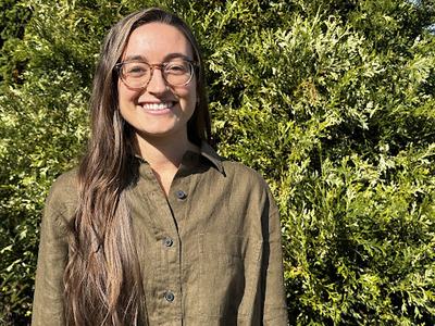 Woman with long hair and glasses standing in front of dense green shrubbery
