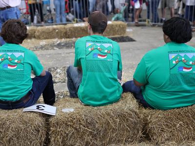 Three people in green "2024 Youth Market Turkey Show" shirts sitting on hay bales at a fair