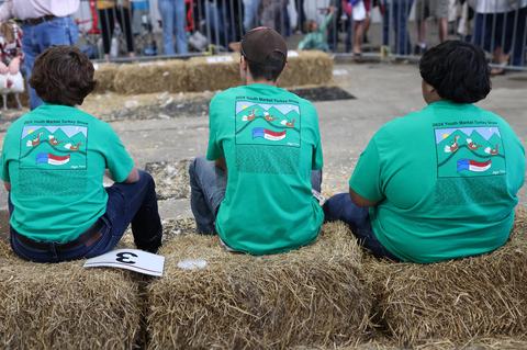 Three people in green "2024 Youth Market Turkey Show" shirts sitting on hay bales at a fair