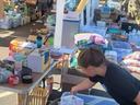 Child placing items into a plastic bag at an outdoor market table filled with goods