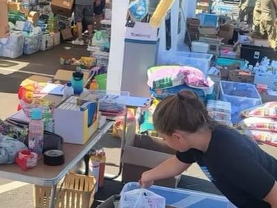 Child placing items into a plastic bag at an outdoor market table filled with goods