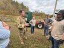 Soldier in camouflage speaking to a group beside a green truck and trailer