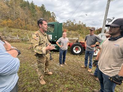 Soldier in camouflage speaking to a group beside a green truck and trailer