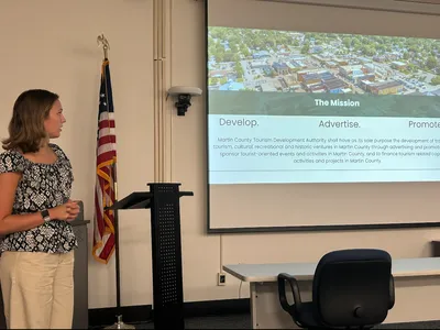A young woman (Emily) in a flower blouse and slacks is standing in a conference room to the left side of a slide show presentation showing an overhead view of a town on top and text below.