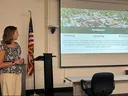 A young woman (Emily) in a flower blouse and slacks is standing in a conference room to the left side of a slide show presentation showing an overhead view of a town on top and text below.