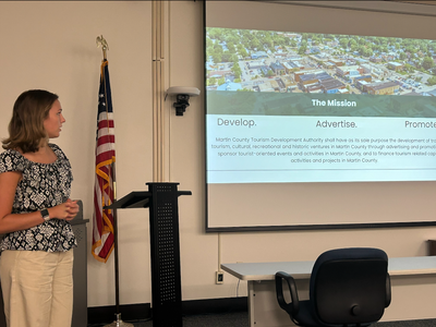A young woman (Emily) in a flower blouse and slacks is standing in a conference room to the left side of a slide show presentation showing an overhead view of a town on top and text below.