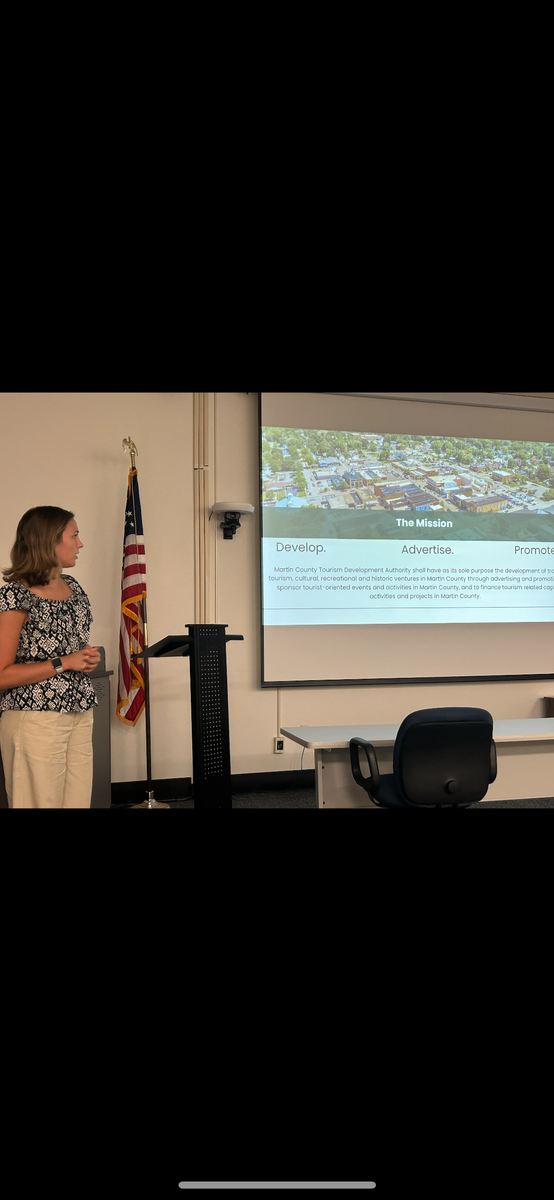 A young woman (Emily) in a flower blouse and slacks is standing in a conference room to the left side of a slide show presentation showing an overhead view of a town on top and text below. 
