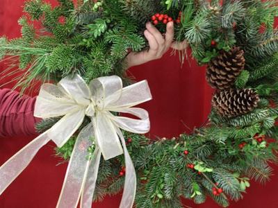 Hand holding evergreen wreath with pinecones, red berries, and white ribbon bow