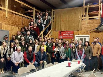 Large group gathered in barn-like hall with sign "NC State Beaufort County Alumni Network"