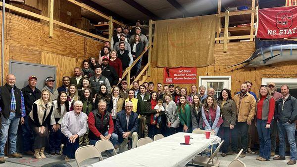 Large group gathered in barn-like hall with sign "NC State Beaufort County Alumni Network"