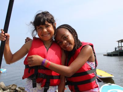 Two girls wearing red life jackets hugging on a lakeshore with paddleboards nearby
