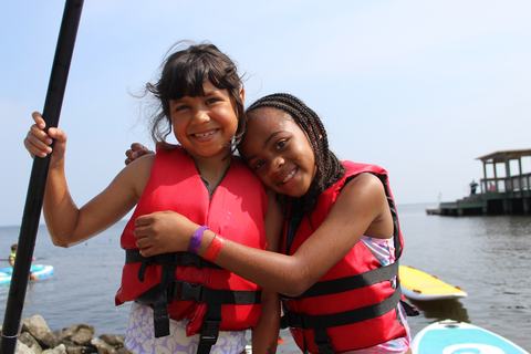 Two girls wearing red life jackets hugging on a lakeshore with paddleboards nearby