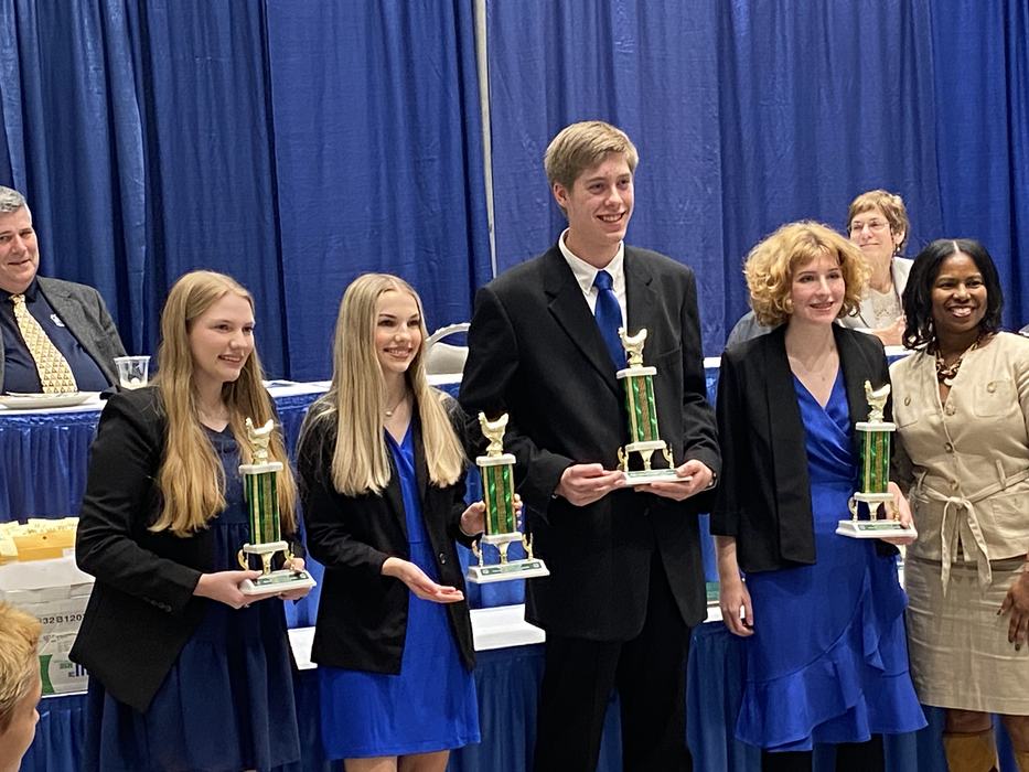 Four people in blue and black holding green trophies on a stage with a blue curtain