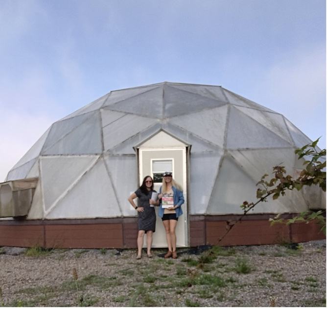 Two people standing at a geodesic dome doorway, one holding a magazine/papers