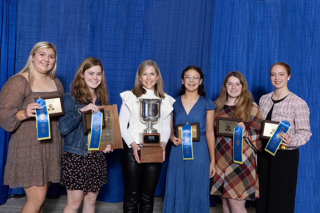 Six women standing in front of blue curtain holding plaques, blue ribbons, and a large trophy.