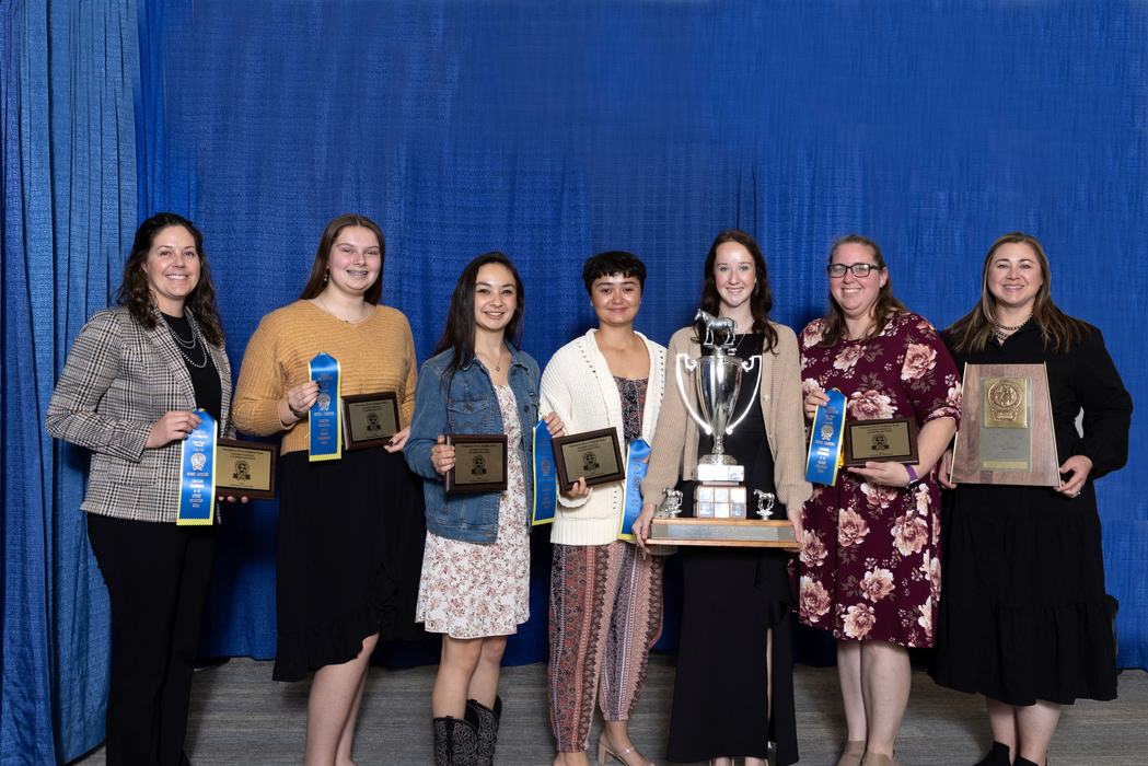 Seven women standing holding plaques, blue ribbons, and a large trophy against blue backdrop