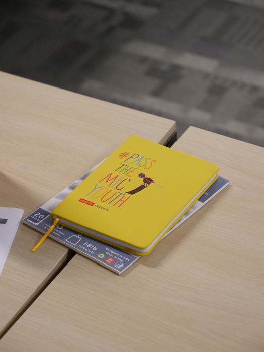 Yellow notebook on table with text "#PASS THE MIC YOUTH" and a microphone graphic.
