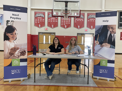 Two SECU staff seated at a table in a school gym with banners reading "Next Payday" and "Help"