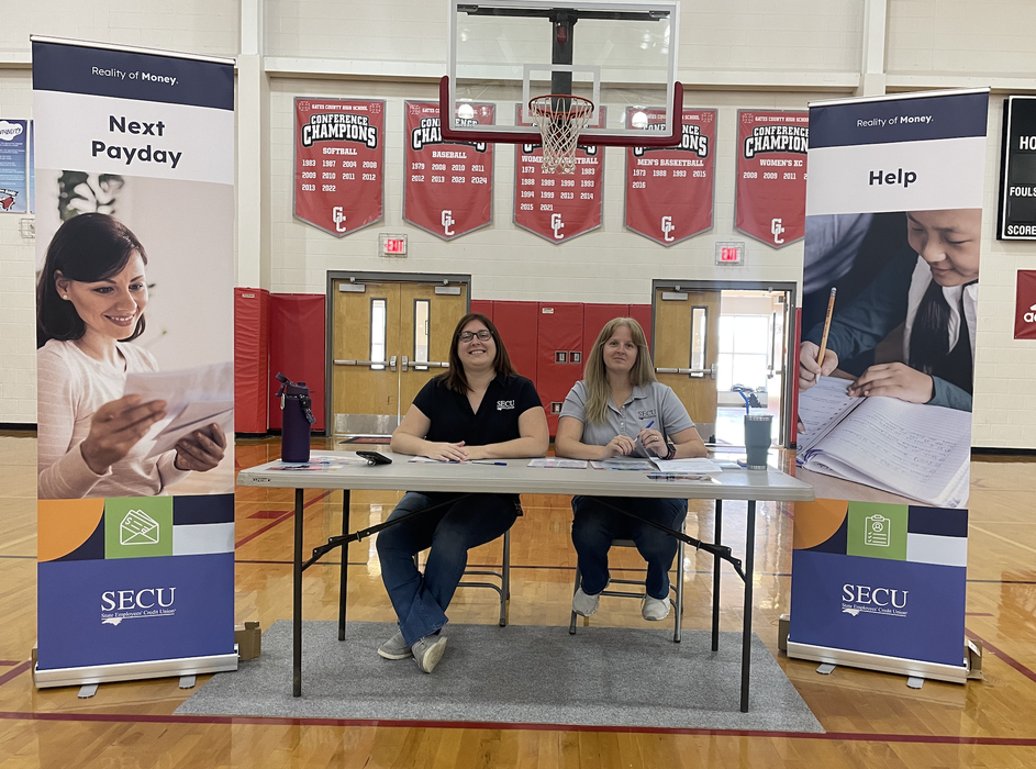 Two SECU staff seated at a table in a school gym with banners reading "Next Payday" and "Help"