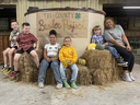 Children and adult sitting on hay bales in front of sign "TRI-COUNTY Broiler Project"