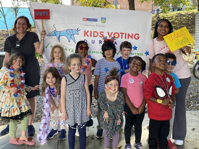 Children and adults in front of a banner reading "KIDS VOTING DURHAM"; signs say "VOTE" and "Voting FUN!"