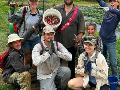 Group of farm workers posing in a field; man holds bucket of harvested okra and sash "Birthday Boy"