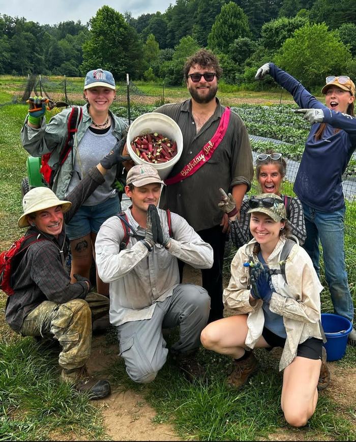 Group of farm workers posing in a field; man holds bucket of harvested okra and sash "Birthday Boy"