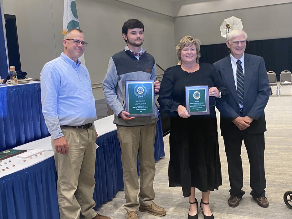 Four people standing in a conference room; two center individuals holding award plaques.