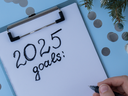 Hand with pen writing "2025 goals:" on clipboard amid decorative rattan balls and silver confetti