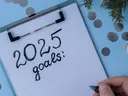 Hand with pen writing "2025 goals:" on clipboard amid decorative rattan balls and silver confetti