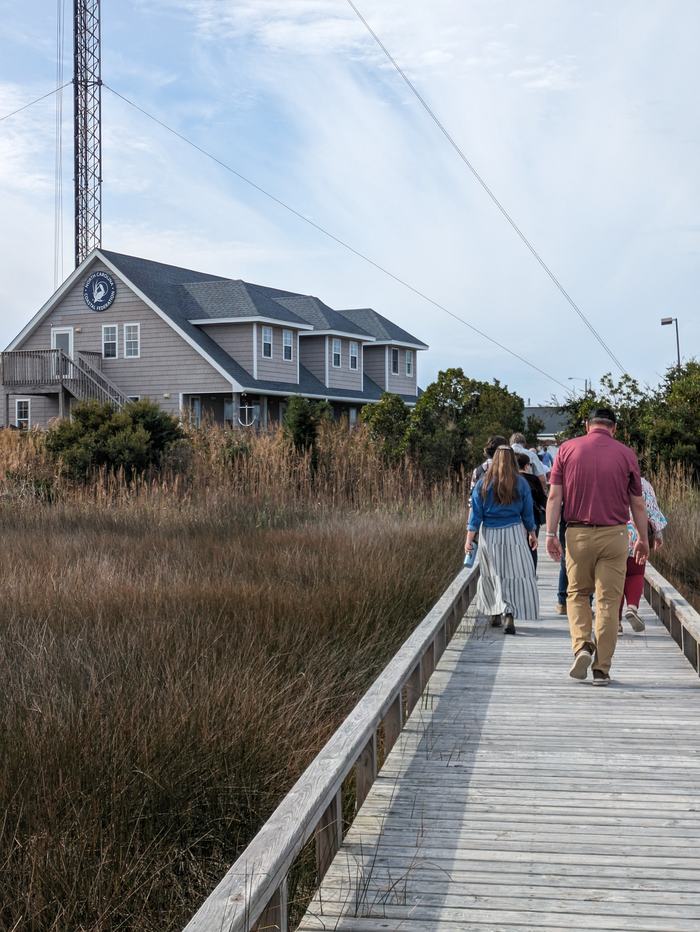 People walking on a wooden boardwalk toward a coastal house beside marshland