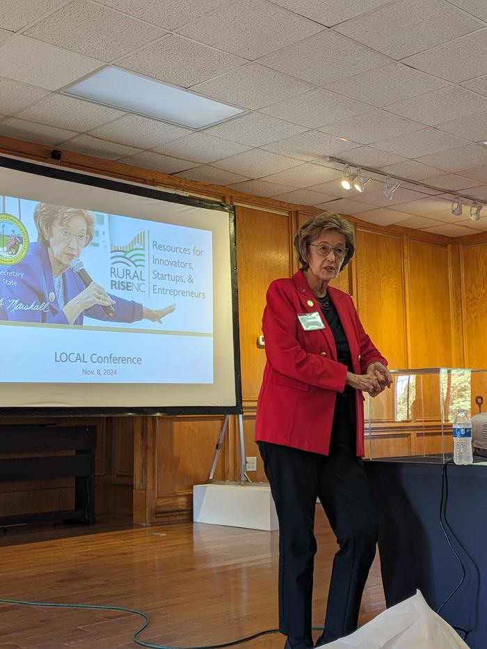 Woman in red jacket speaking beside screen: "RURAL RISE NC" and "LOCAL Conf. Nov 8, 2024"