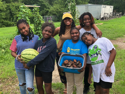 Six children standing outdoors; one holds a watermelon, one holds a bucket of potatoes; shirt reads "Henderson Collegiate"