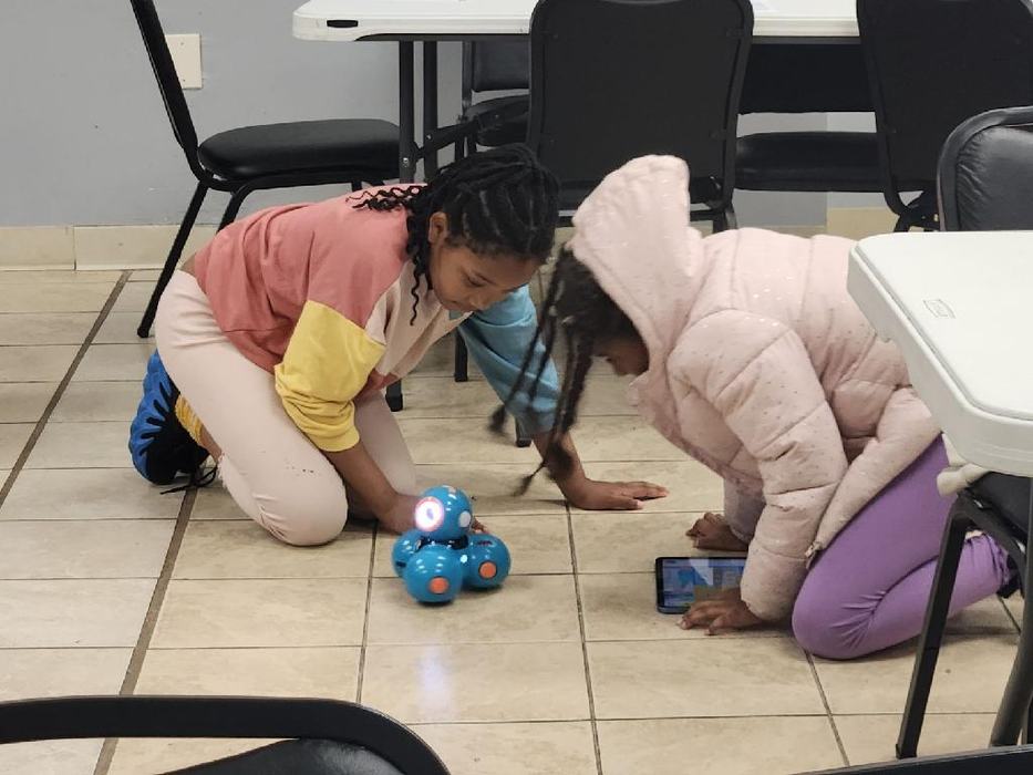 Two students watch as a blue robot moves across the floor.