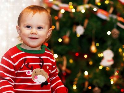 Toddler in red striped sweater with reindeer motif standing in front of decorated Christmas tree.