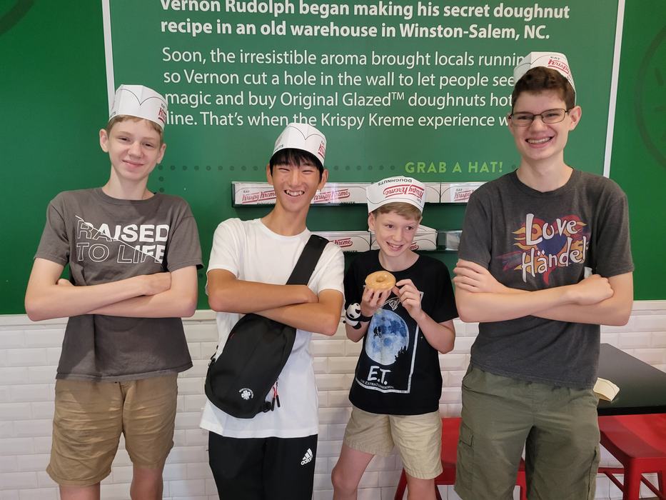 Four boys (three brothers and their exchange delegate from Japan) stand together in a Krispy Kreme donut shop wearing paper hats from the shop. They are smiling and laughing. The smallest boy is holding a glazed doughnut.