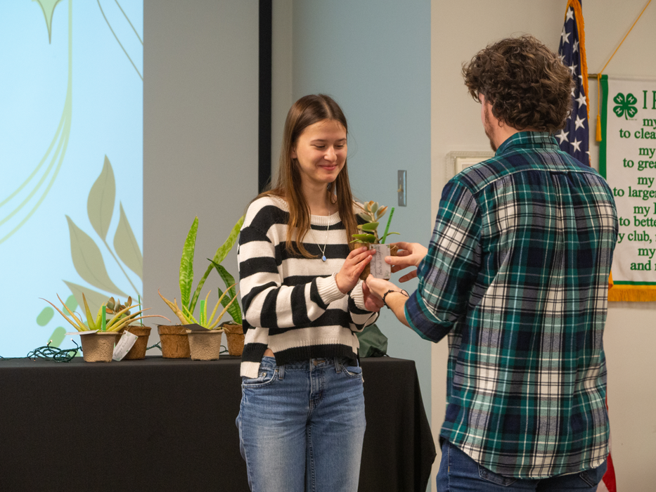 Young woman receives a small potted plant from a person in a plaid shirt