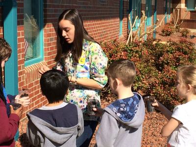 Adult showing seedlings to five children holding clear cups of soil outside a brick school
