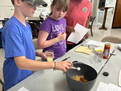 Three children at a table mixing ingredients; boy stirring a bowl while others hold paper and cup.