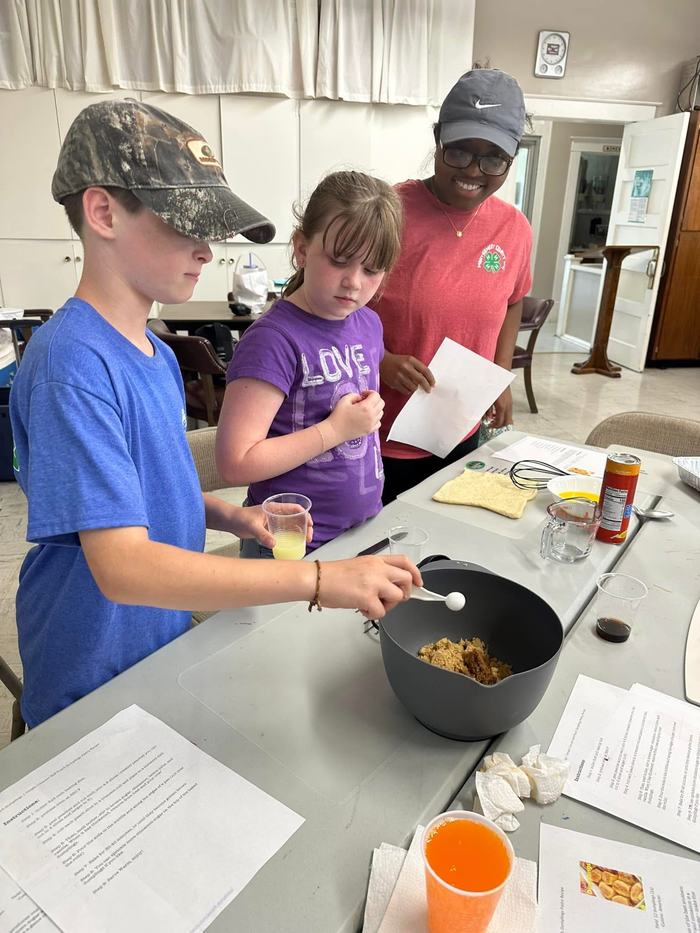 Three children at a table mixing ingredients; boy stirring a bowl while others hold paper and cup.